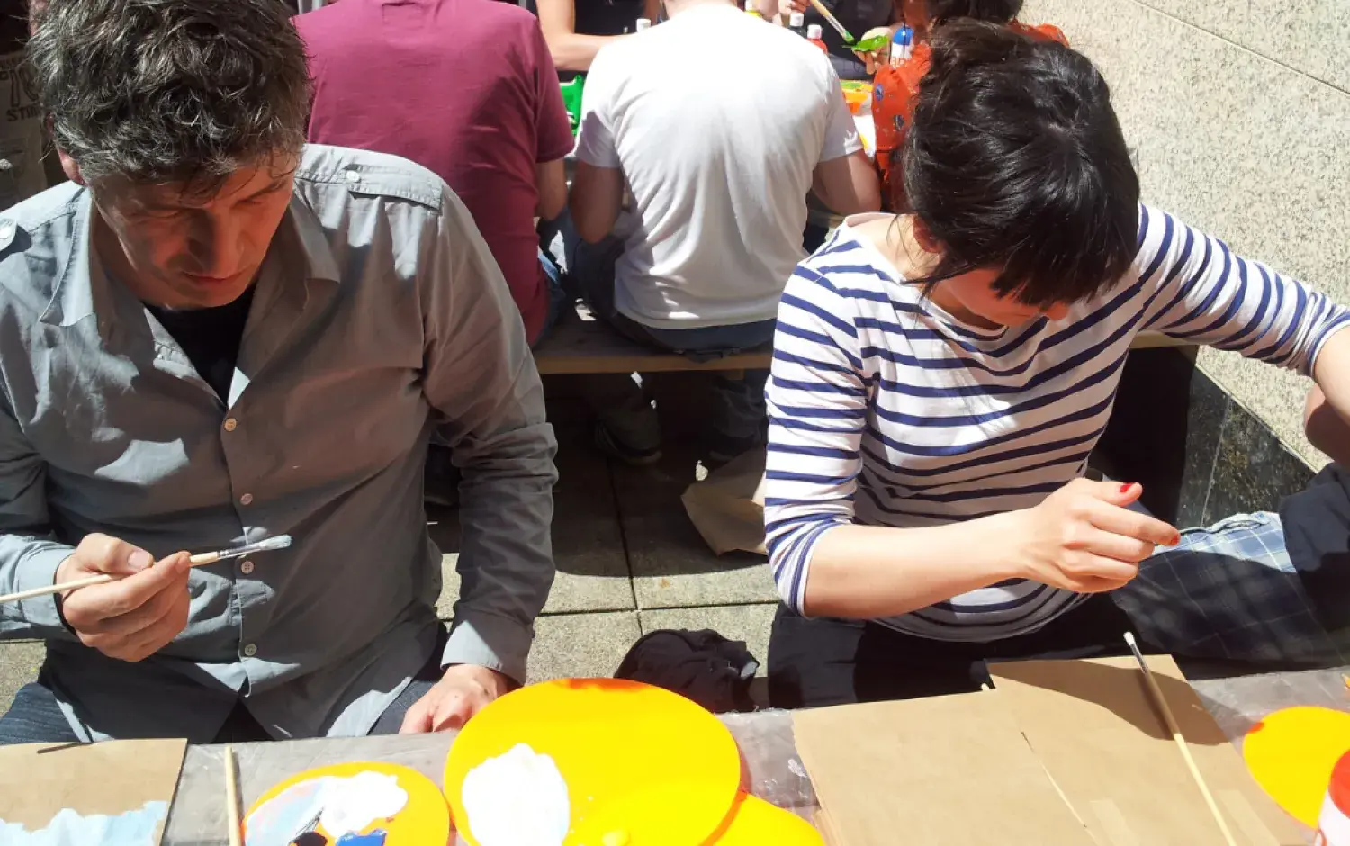 Two people seated at a table outdoors, painting small objects with brushes; bright orange plates and paint supplies are on the table.