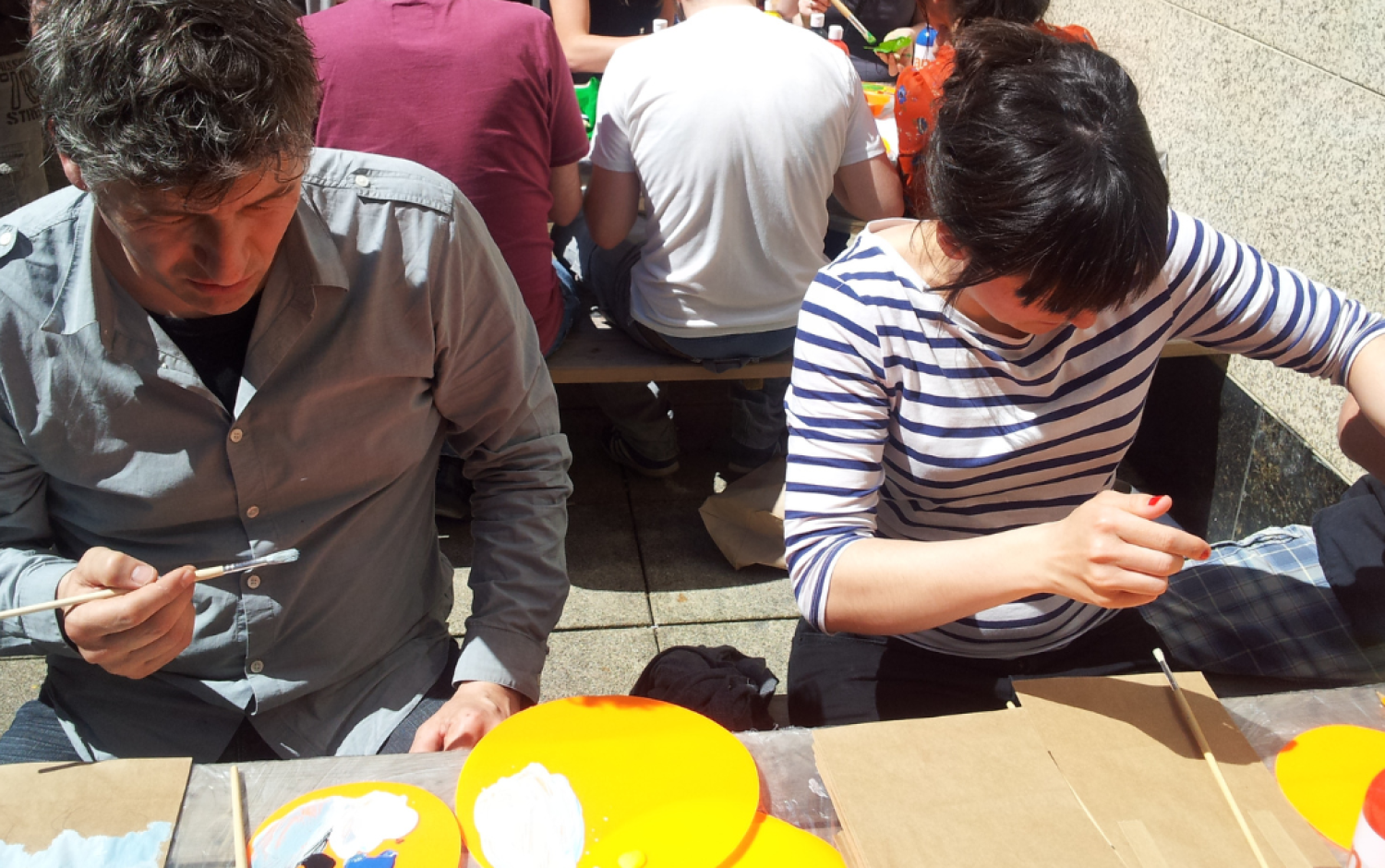 Two people seated at a table outdoors, painting small objects with brushes; bright orange plates and paint supplies are on the table.