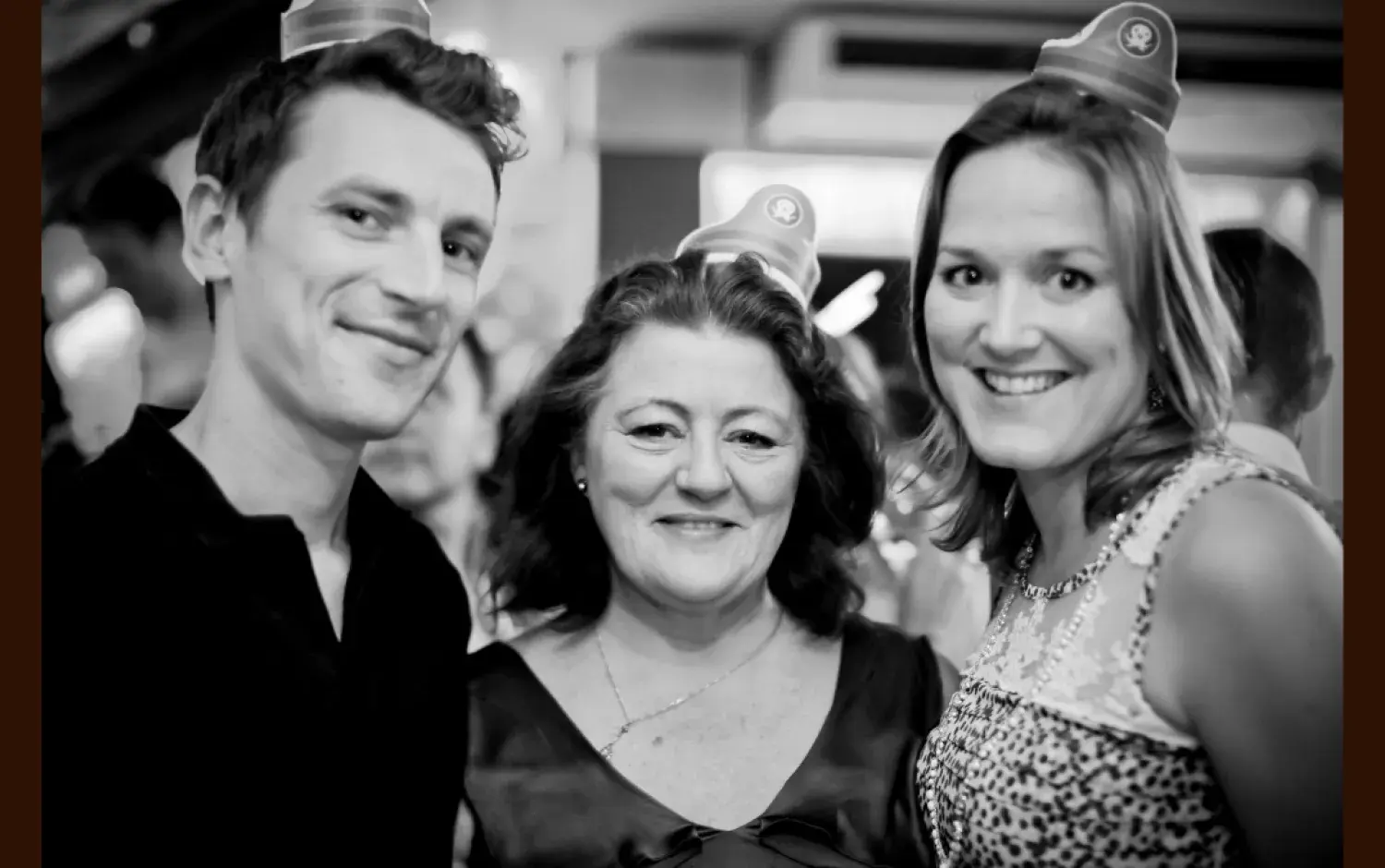 Black-and-white photo of three people wearing small party hats, standing close together at an indoor event.