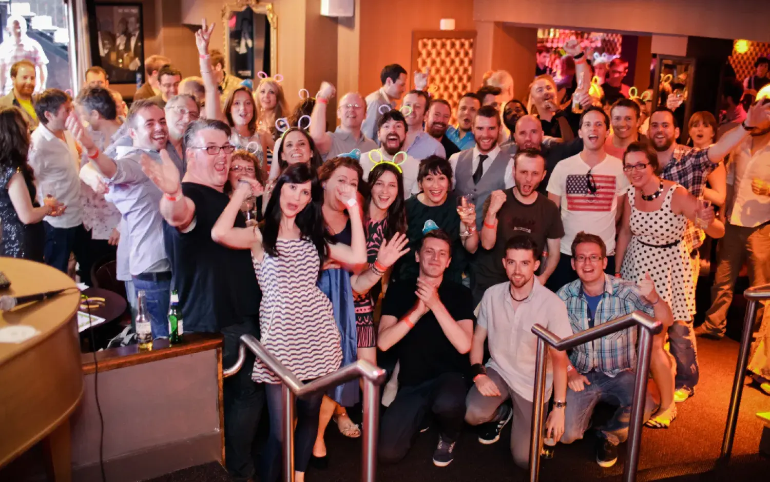 Group photo of people posing in a lively indoor venue, many wearing novelty headbands.