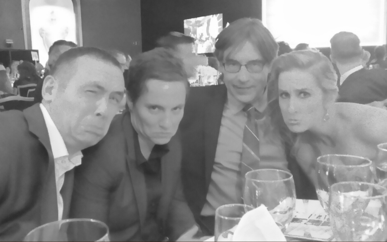 Black-and-white photo of four people seated at a table during a formal dinner event with glasses and menus visible.
