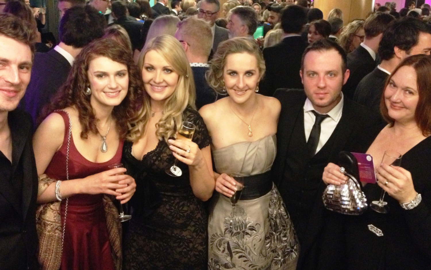Group of people dressed in formal attire holding champagne glasses at a crowded awards event.