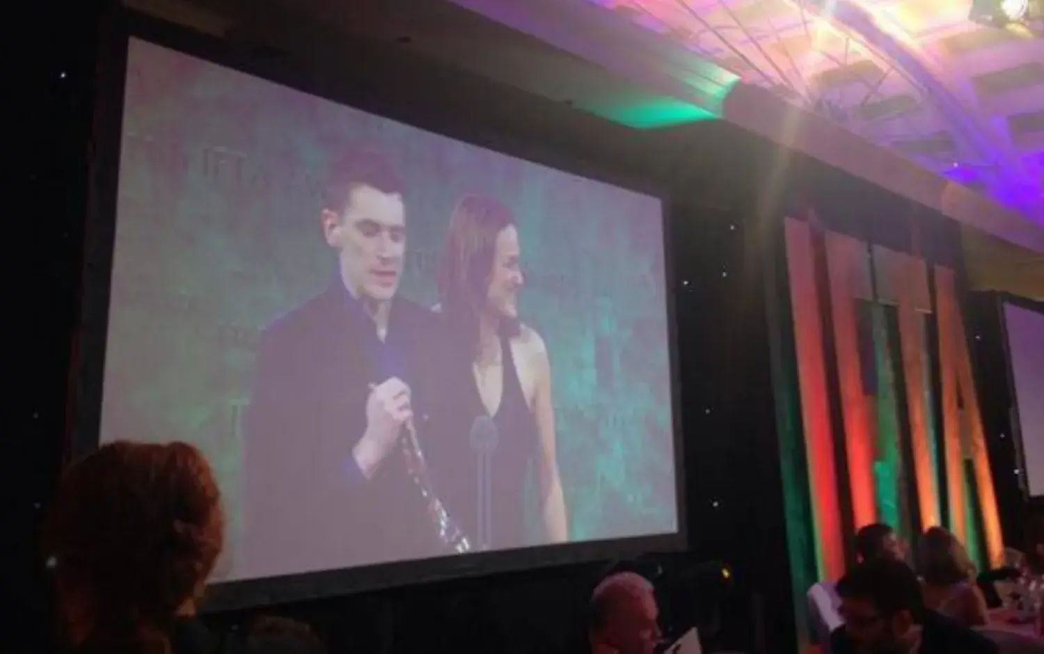 Projected image of two people on stage at an awards ceremony, with audience seated in front.