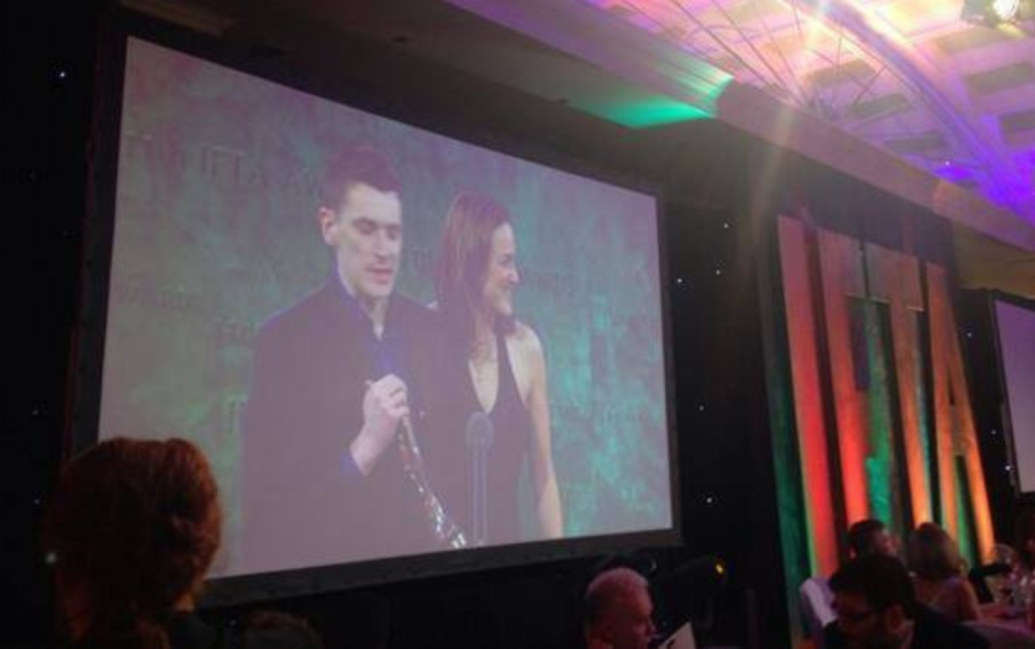 Projected image of two people on stage at an awards ceremony, with audience seated in front.