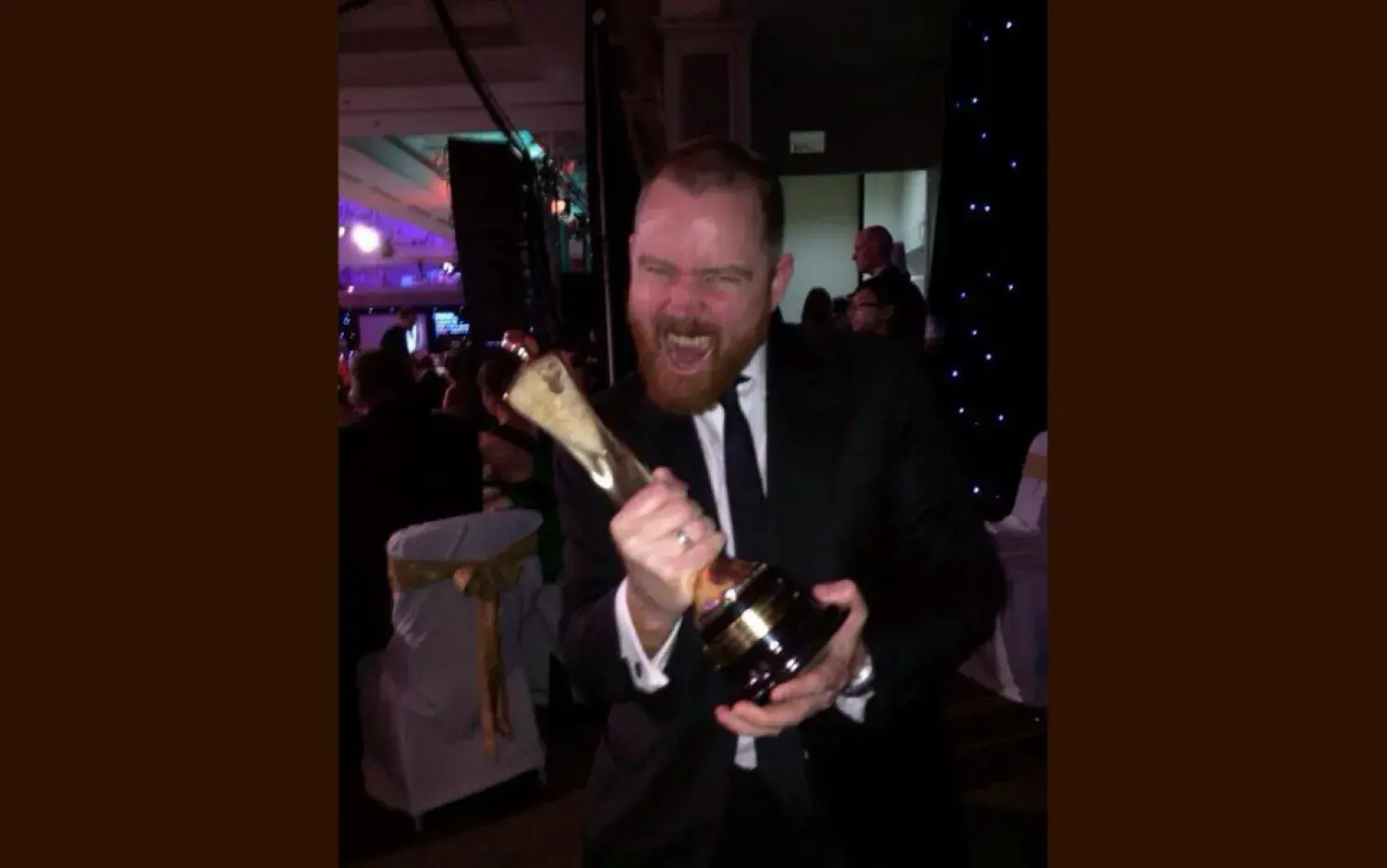 Person in formal attire holding a tall award trophy at an event with decorated tables.