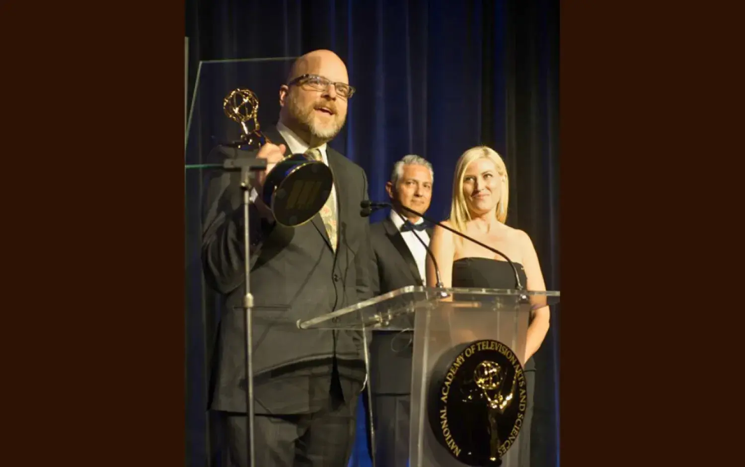 Person speaking at podium holding an Emmy Award during a formal ceremony.