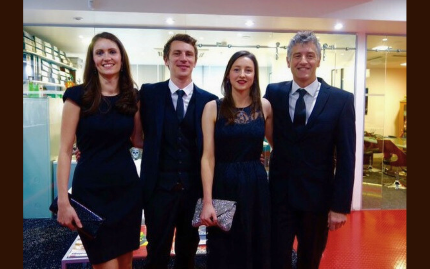 Four people dressed in black formal wear posing in a brightly lit room.