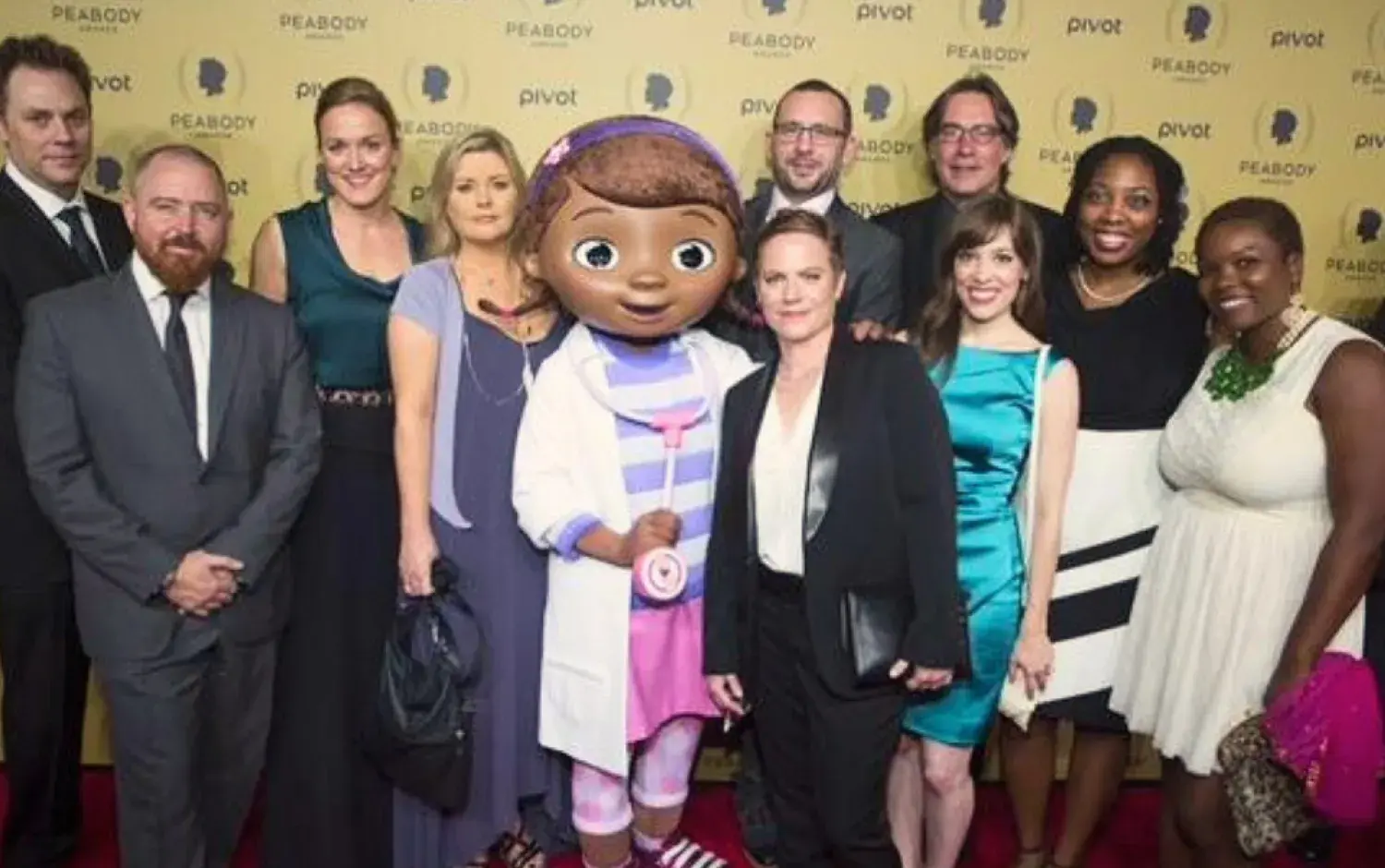 Large group posing on a Peabody Awards red carpet with a character mascot in the center.