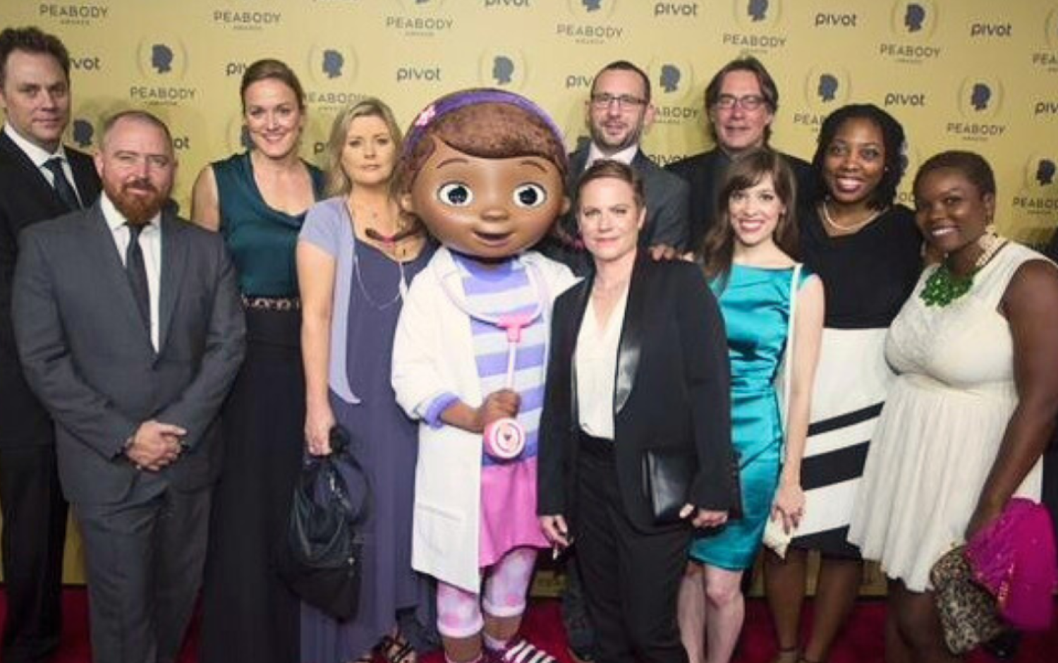 Large group posing on a Peabody Awards red carpet with a character mascot in the center.