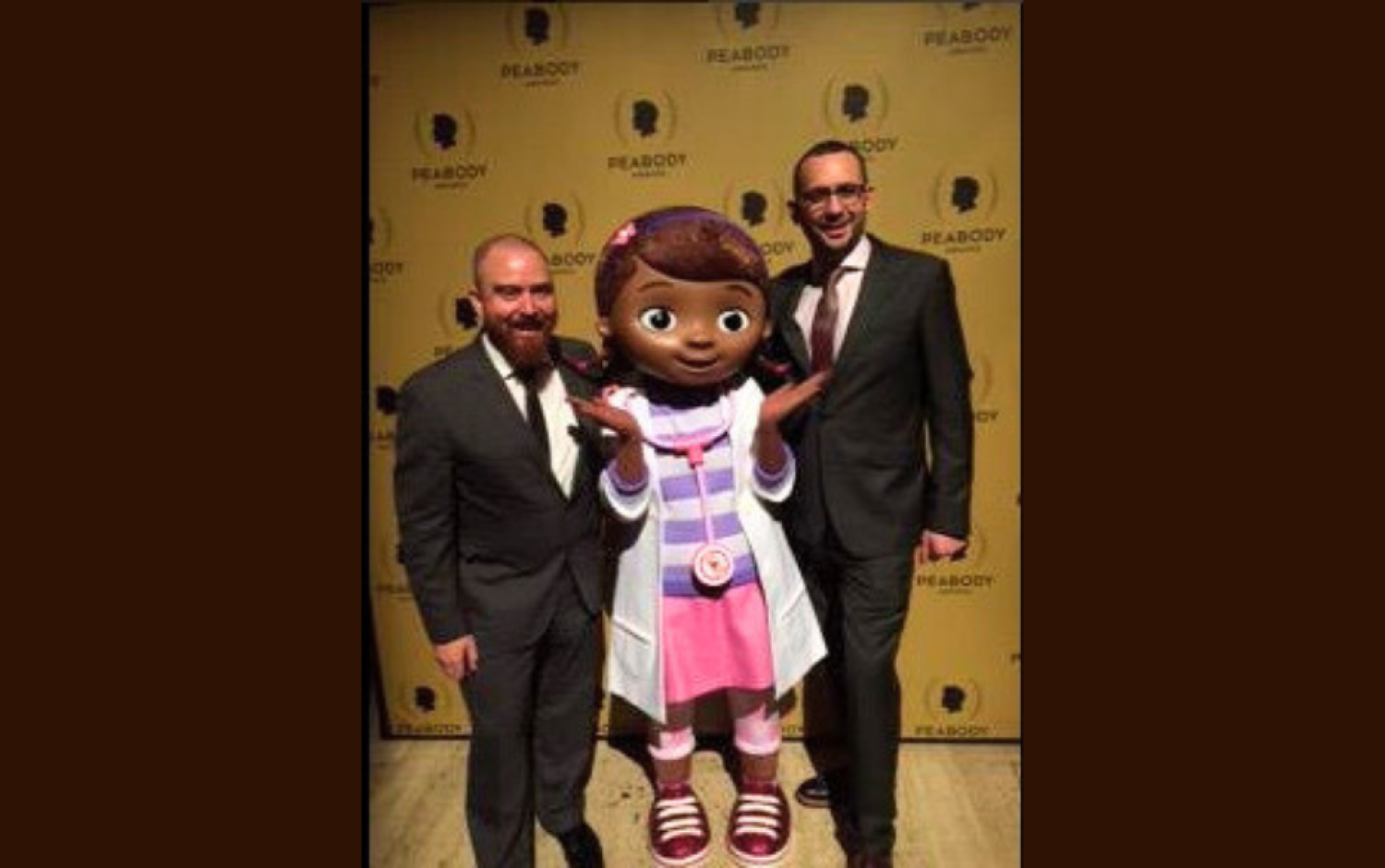 Two people posing with a Doc McStuffins mascot on Peabody Awards backdrop.