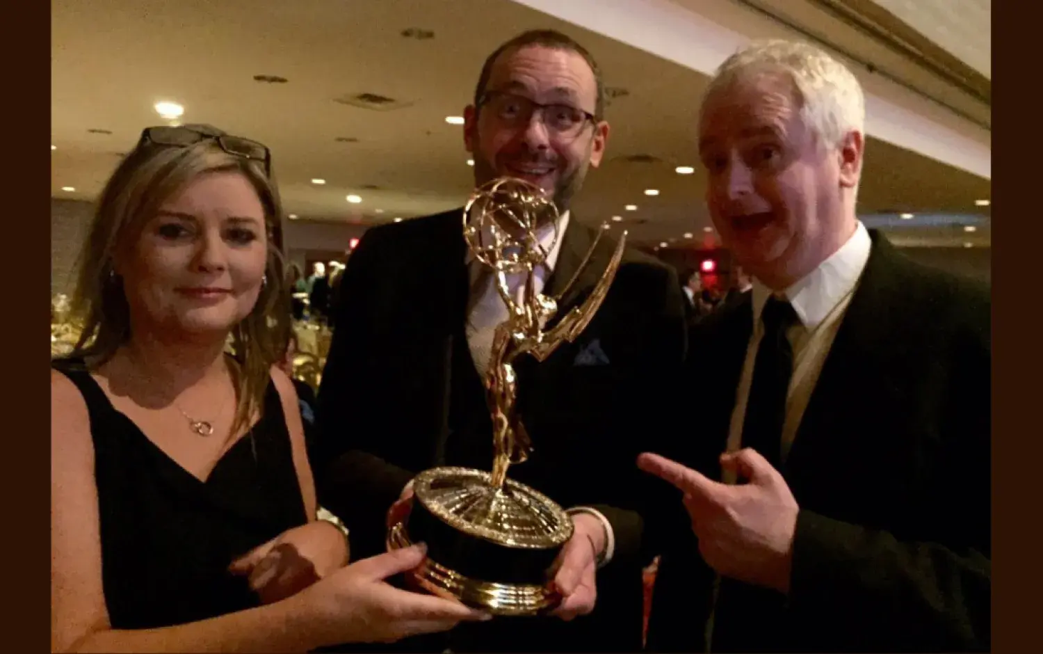 Three people holding an Emmy Award trophy at a formal event.