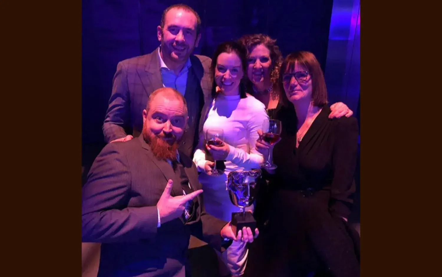 Group seated in a dark venue holding drinks and a BAFTA award trophy..