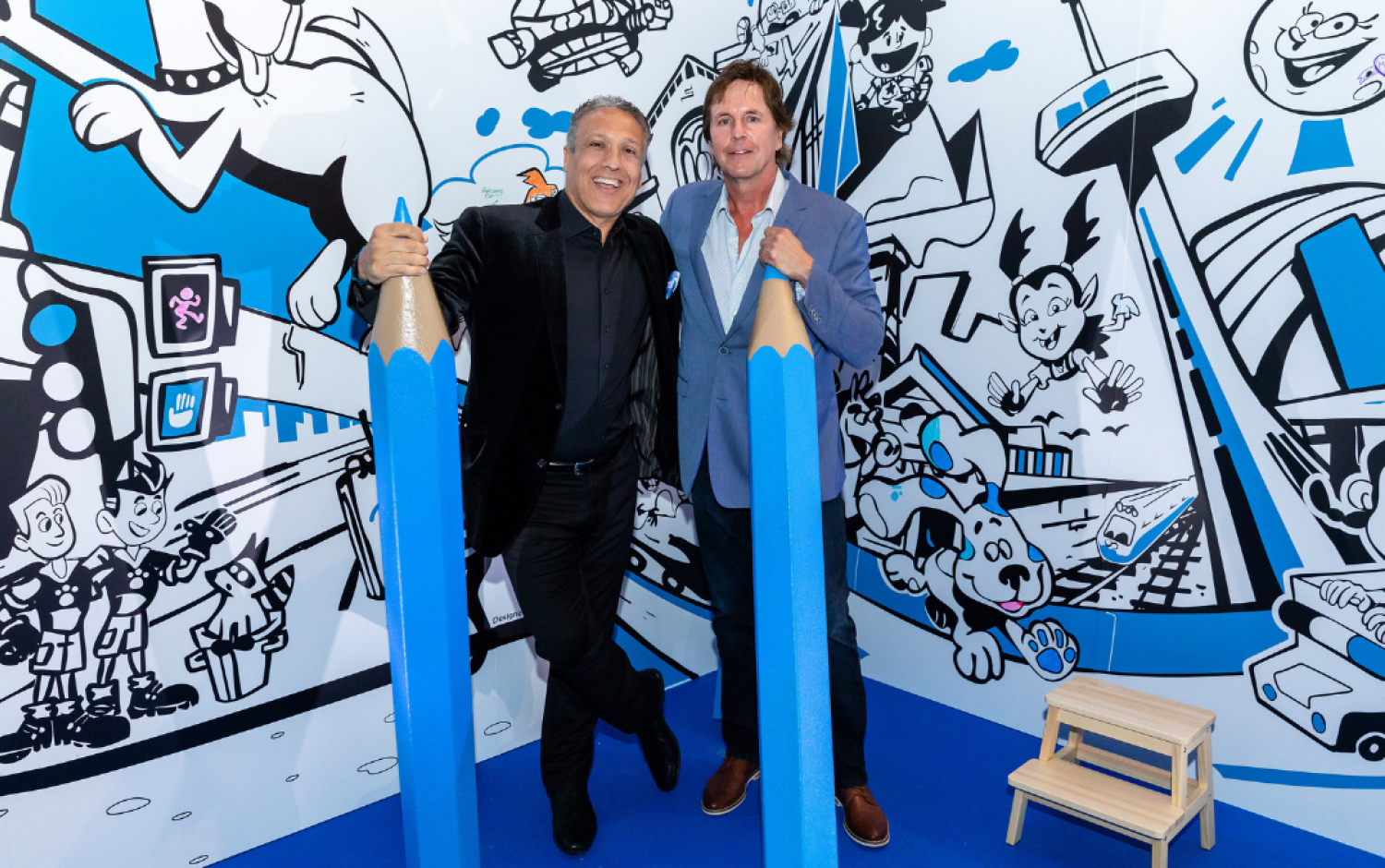 Two people posing with oversized blue pencils in front of a cartoon-themed wall mural.