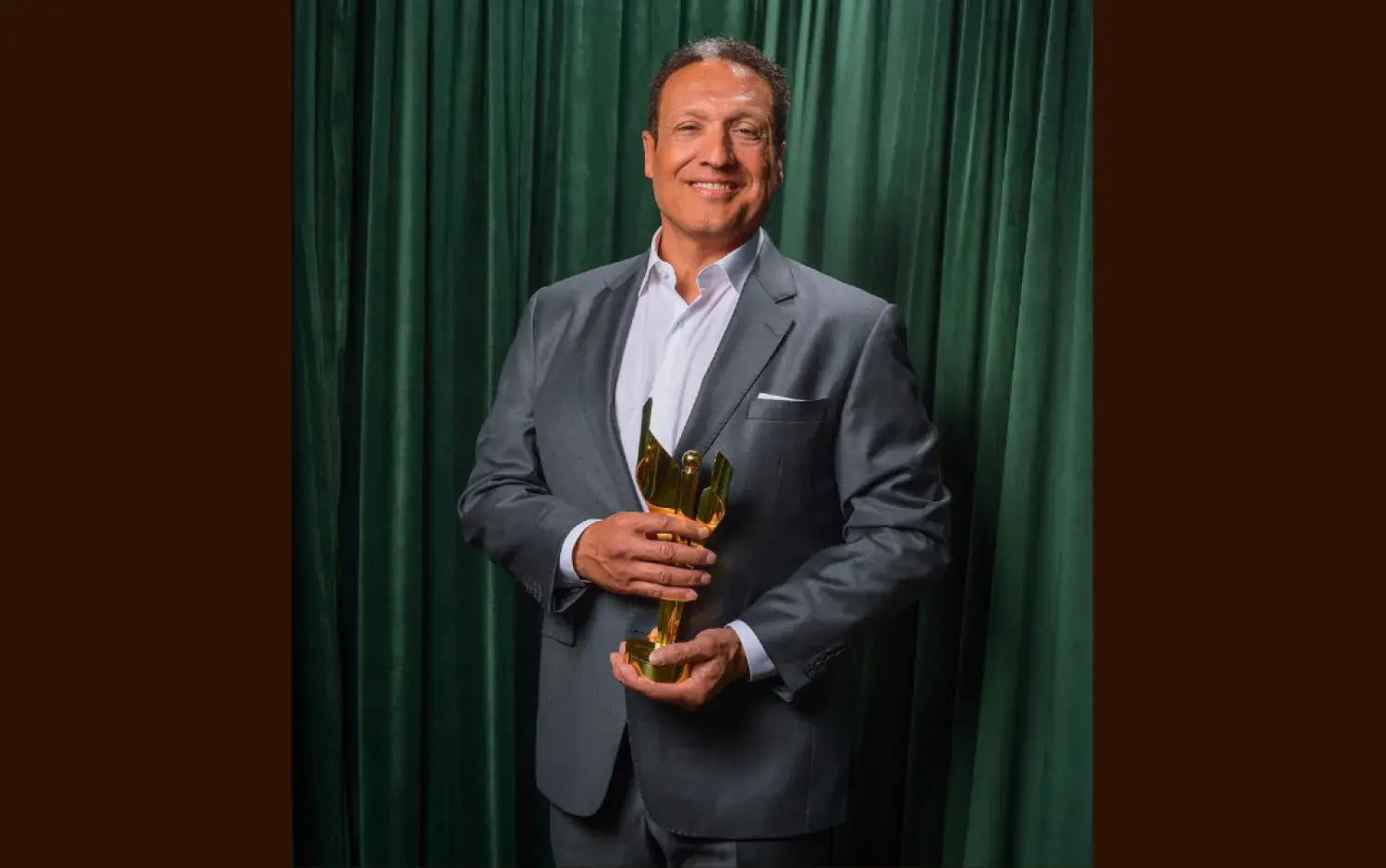 Person in a gray suit holding a golden award trophy against a green curtain backdrop.