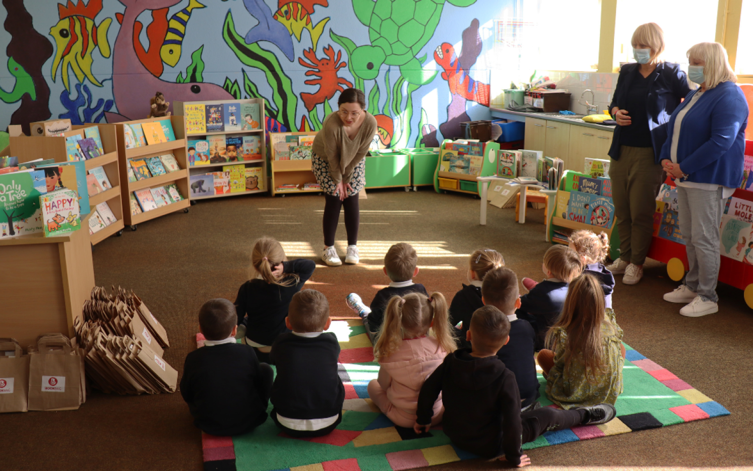 Group of young children seated on a colorful rug listening to an adult in a classroom decorated with books and wall art.