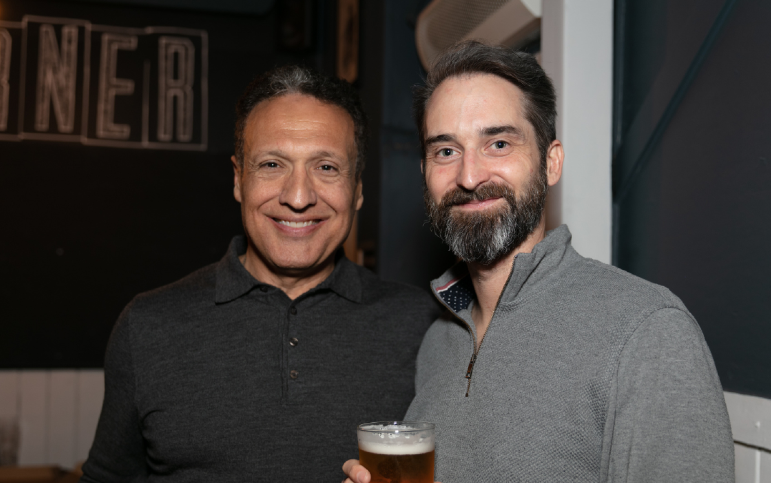 Two people standing close together in a dimly lit bar, one holding a pint of beer.