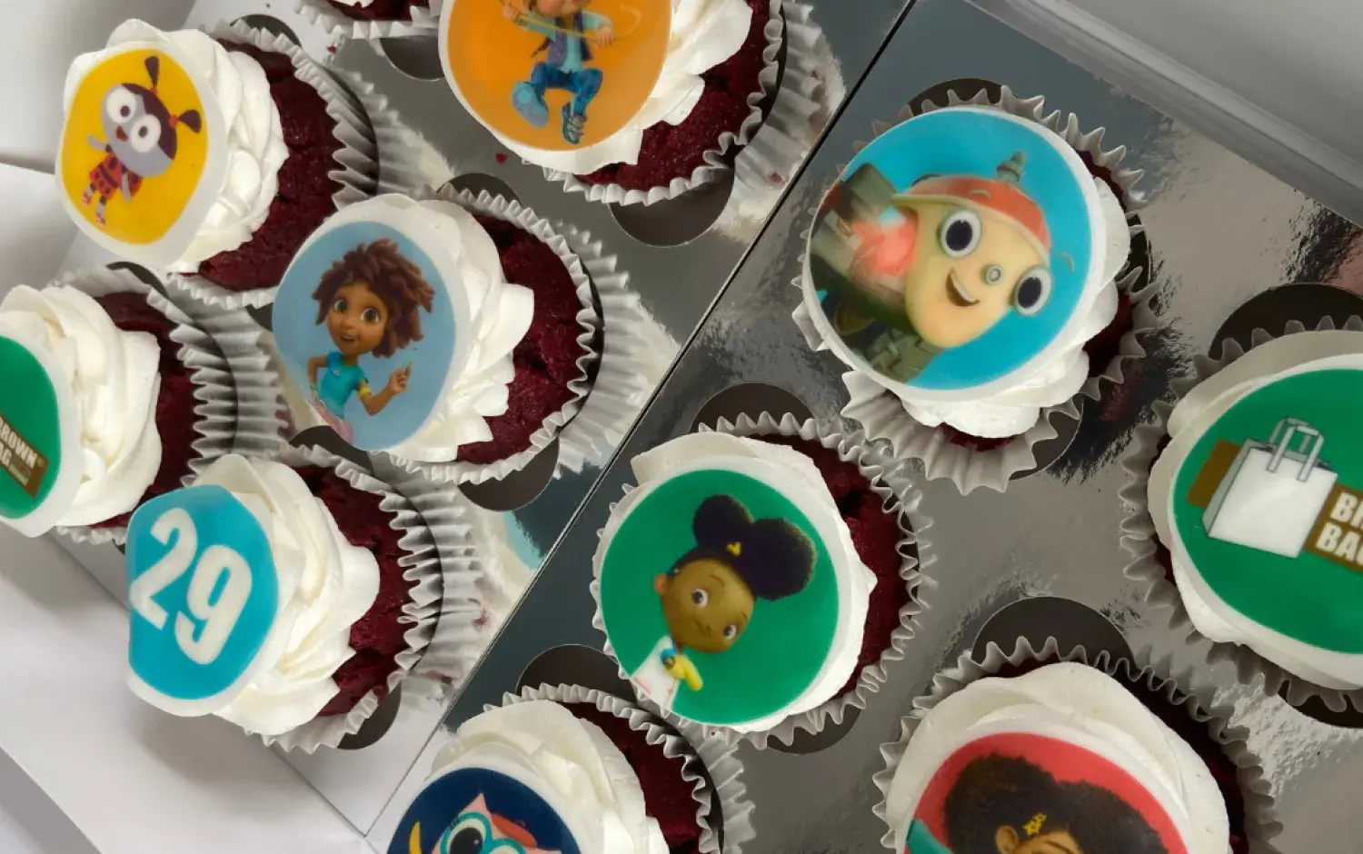 Box of cupcakes decorated with edible images of characters, logos, and the number 29.