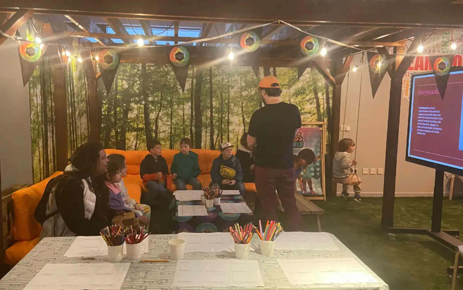Children seated on an orange couch listening to a presenter in a decorated room with art supplies and a large screen.