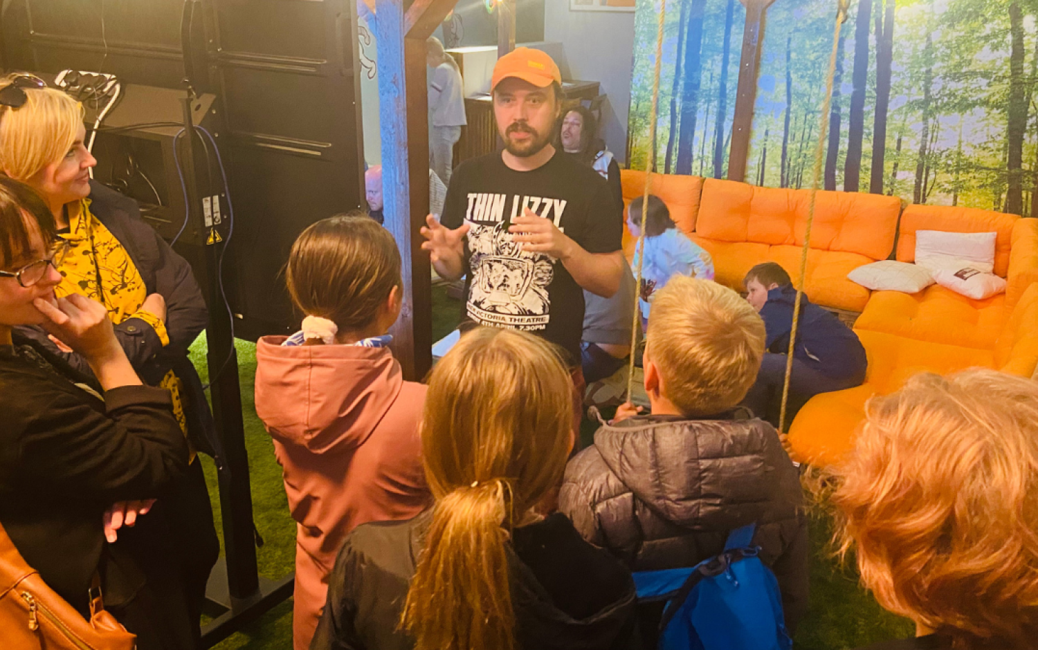 Group gathered around a person speaking in a colorful room with an orange couch and forest-themed wall decor.