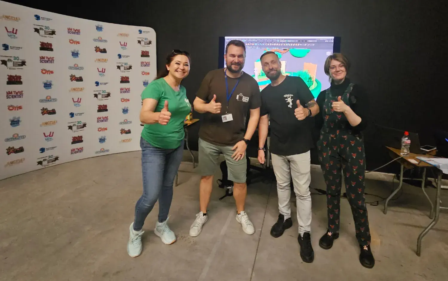 Four people posing in front of a branded backdrop and screen, giving thumbs-up gestures.