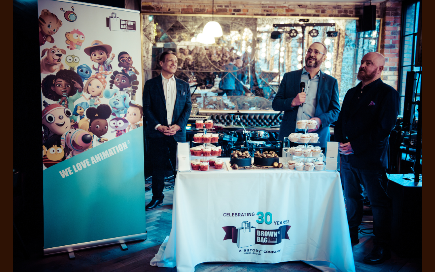 Three people standing behind a table with cupcakes and a banner reading “We Love Animation” at a company celebration.