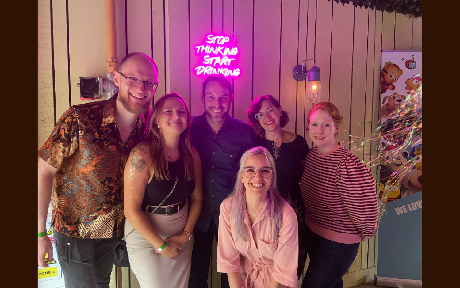 Group posing indoors under a neon sign that reads “Stop Thinking Start Drinking,” with festive lights and a banner featuring animated characters.
