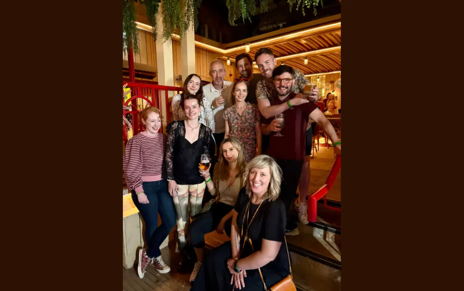 Group gathered together in a warmly lit venue with drinks in hand and greenery hanging overhead.