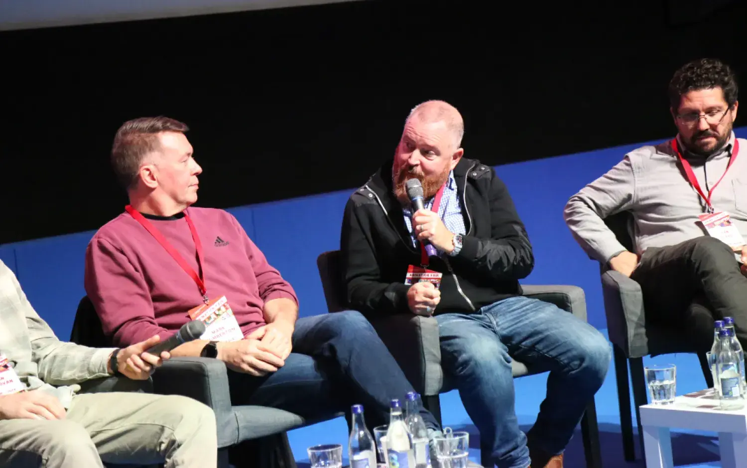 Panel discussion with speakers seated on stage holding microphones, water glasses on a table in front.