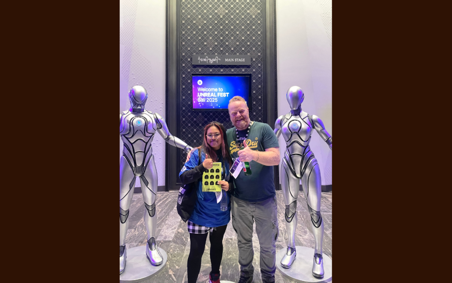 Two people standing between silver humanoid statues holding an award in front of a screen reading “Welcome to Unreal Fest 2025.”