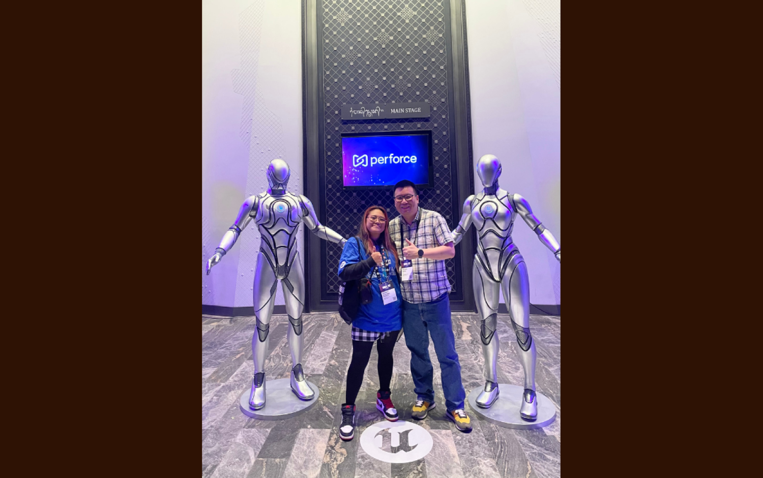Two people posing between two silver humanoid statues in front of a Perforce sign at an event venue.