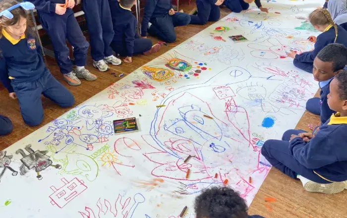 Schoolchildren seated and kneeling on the floor around a large sheet of paper covered in colourful drawings, with crayons and pencils scattered across the surface.