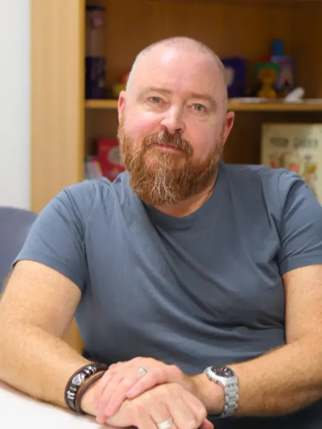 A portrait photo of Darragh O'Connell seated with hands in front of him. A bookshelf in the background.
