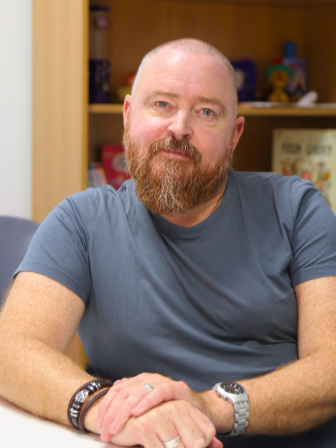 A portrait photo of Darragh O'Connell seated with hands in front of him. A bookshelf in the background.